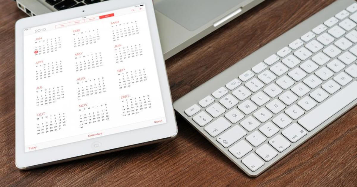 A tablet showing a calendar next to a keyboard on a wooden desk