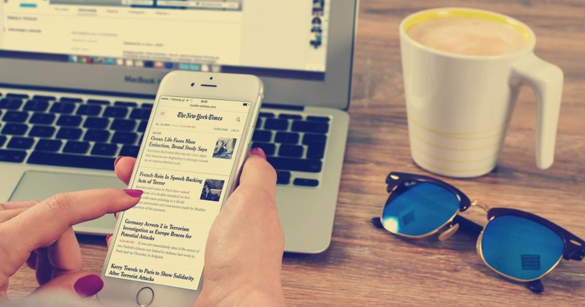 A hand scrolling a news feed on a phone beside a laptop and coffee