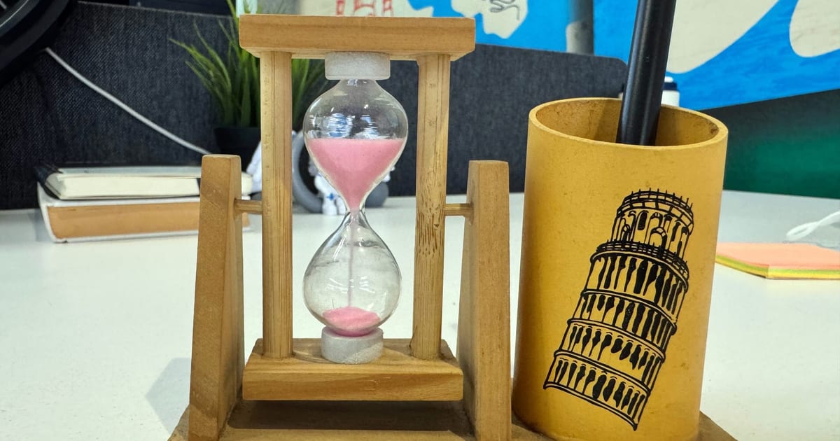 An hourglass with pink sand on a desk beside a pen holder
